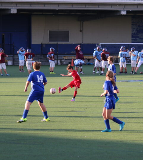 A kid in a red jersey kicks a soccer ball while two kids in blue jerseys defend