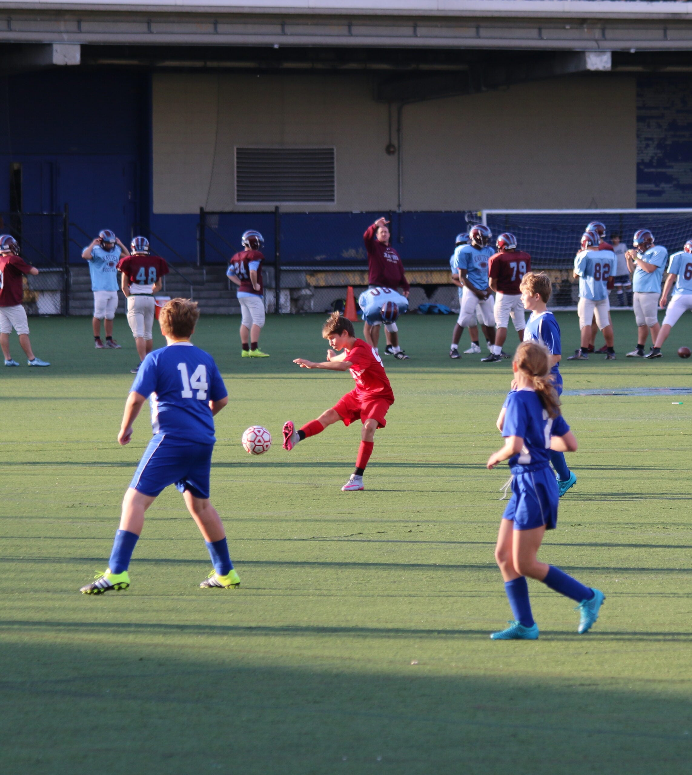 A kid in a red jersey kicks a soccer ball while two kids in blue jerseys defend