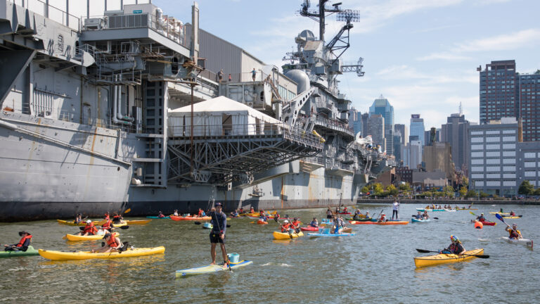 Kayakers and stand-up paddleboarders paddle in the water at Pier 84, with the Intrepid Museum air craft carrier towering above