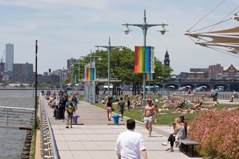 A large gathering of park visitors sit on the green lawn at Pier 45 sunbathing and admiring the views of the Hudson River