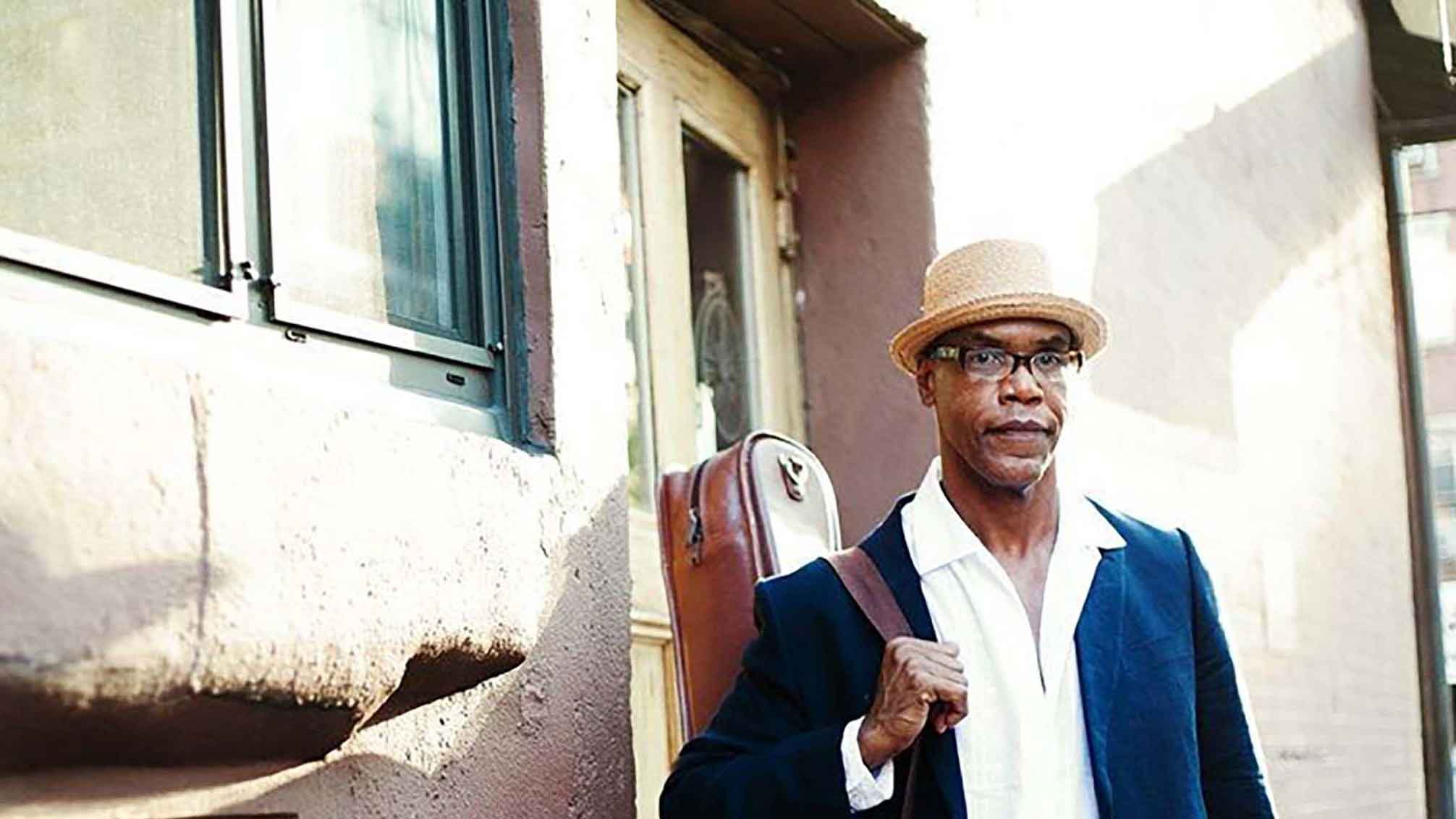 Jazz musician Ed Cherry stands on a balcony with his leather cased guitar and hat