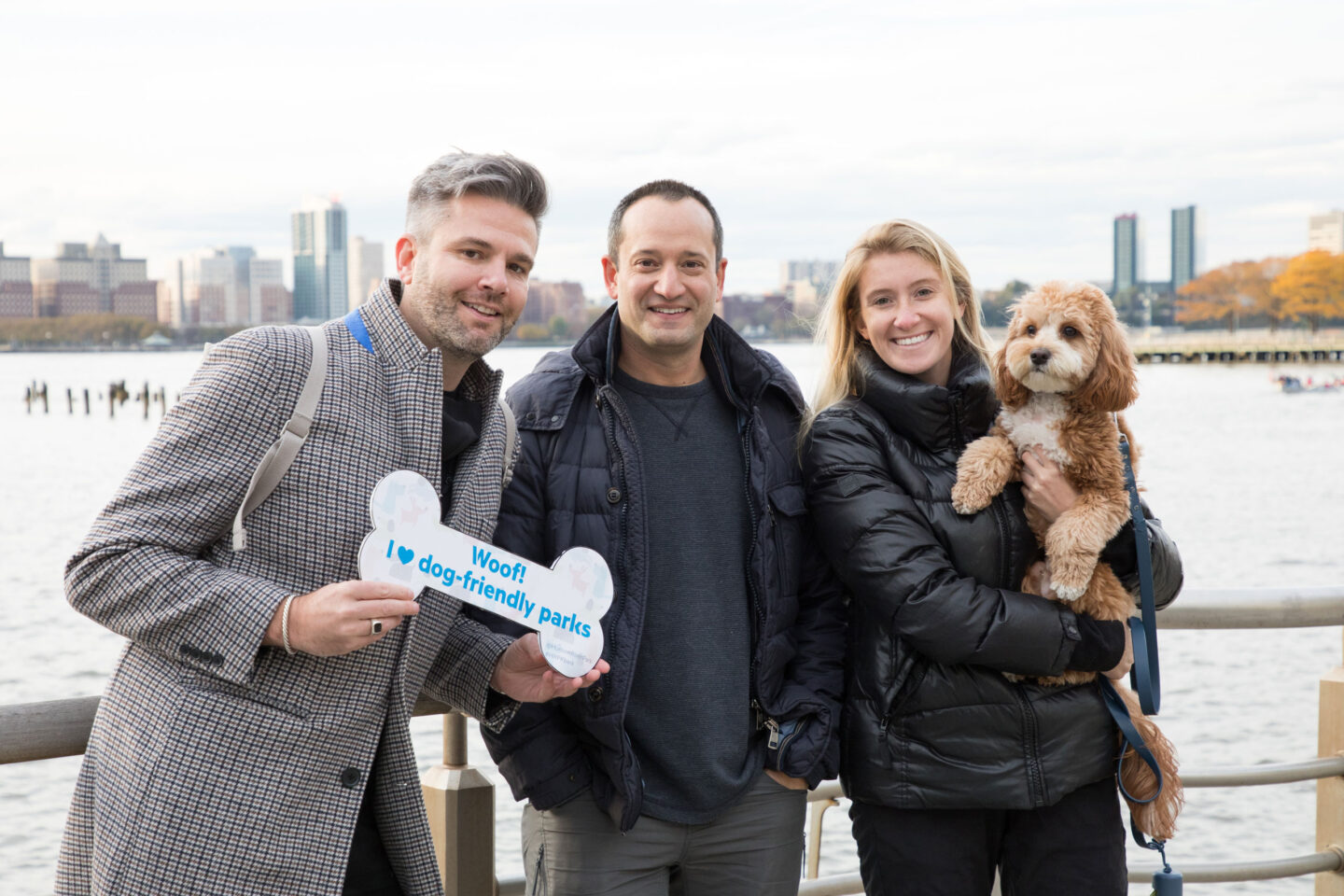 Three people posing with a dog. One person is holding a bone-shaped sign that says, "Woof! I love dog-friendly parks"