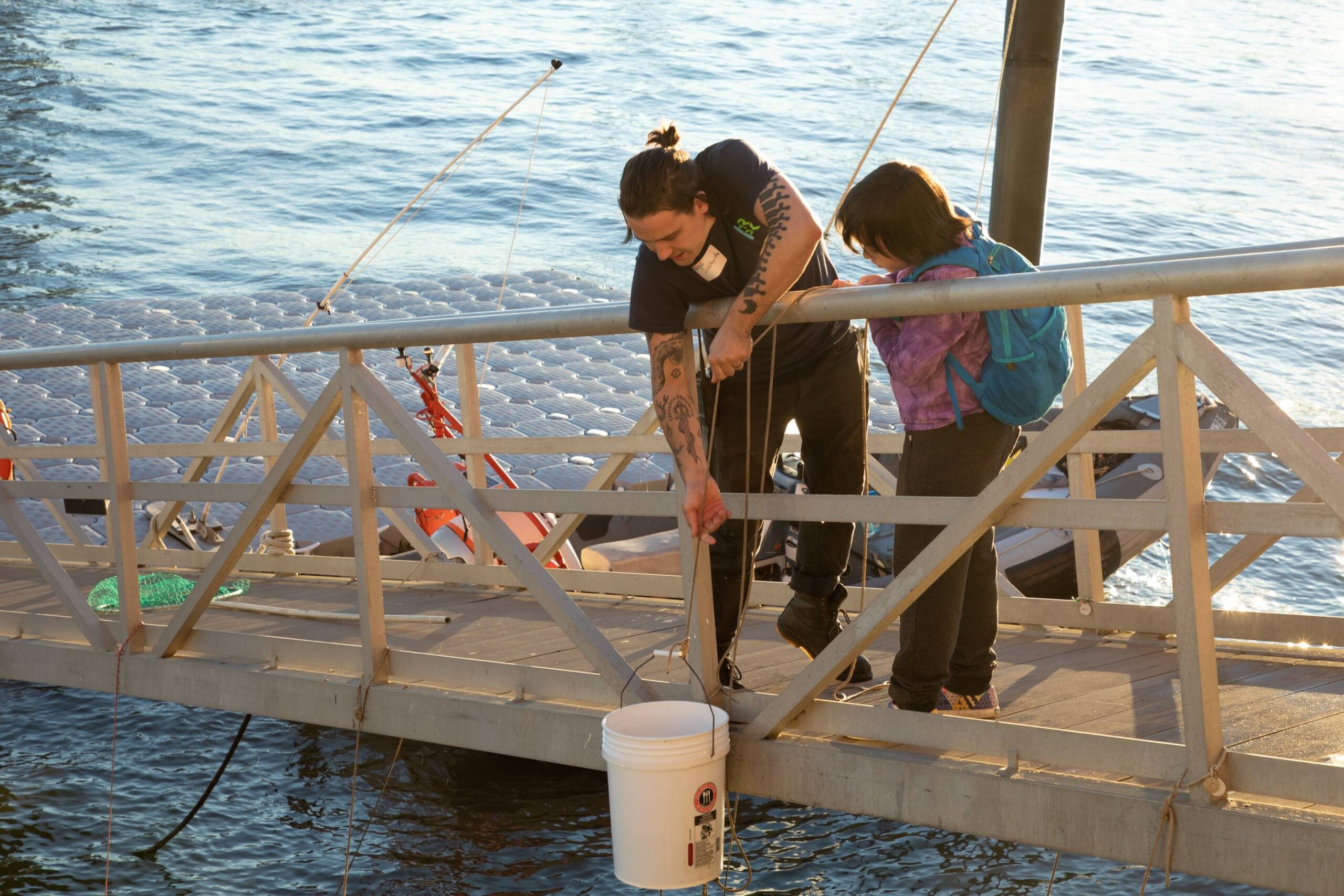 An HRPK River Project staff member lowers a bucket with fish from a dock into the River while a child looks on