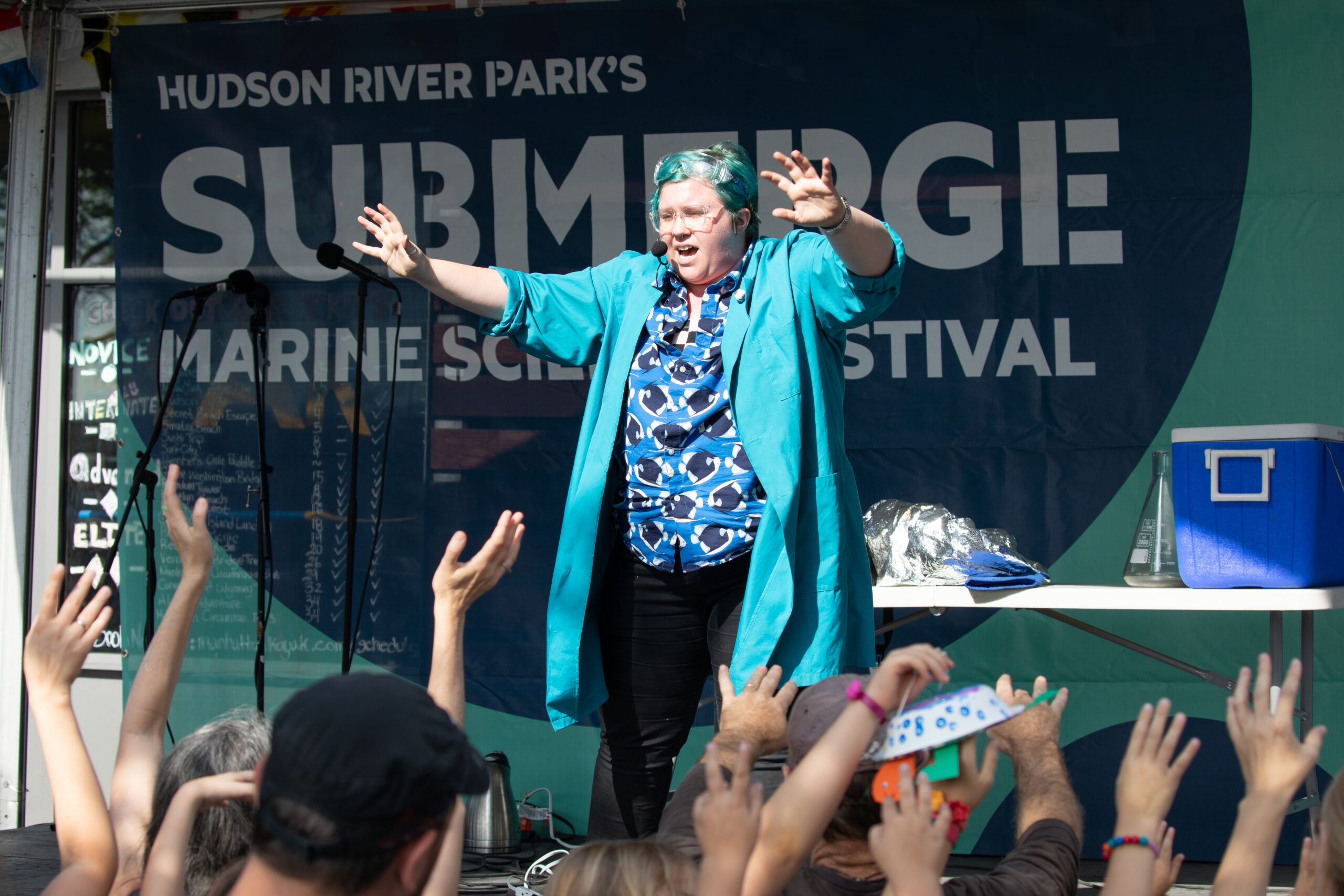 Person with aqua hair and lab coat holds hands out before crowd doing the same
