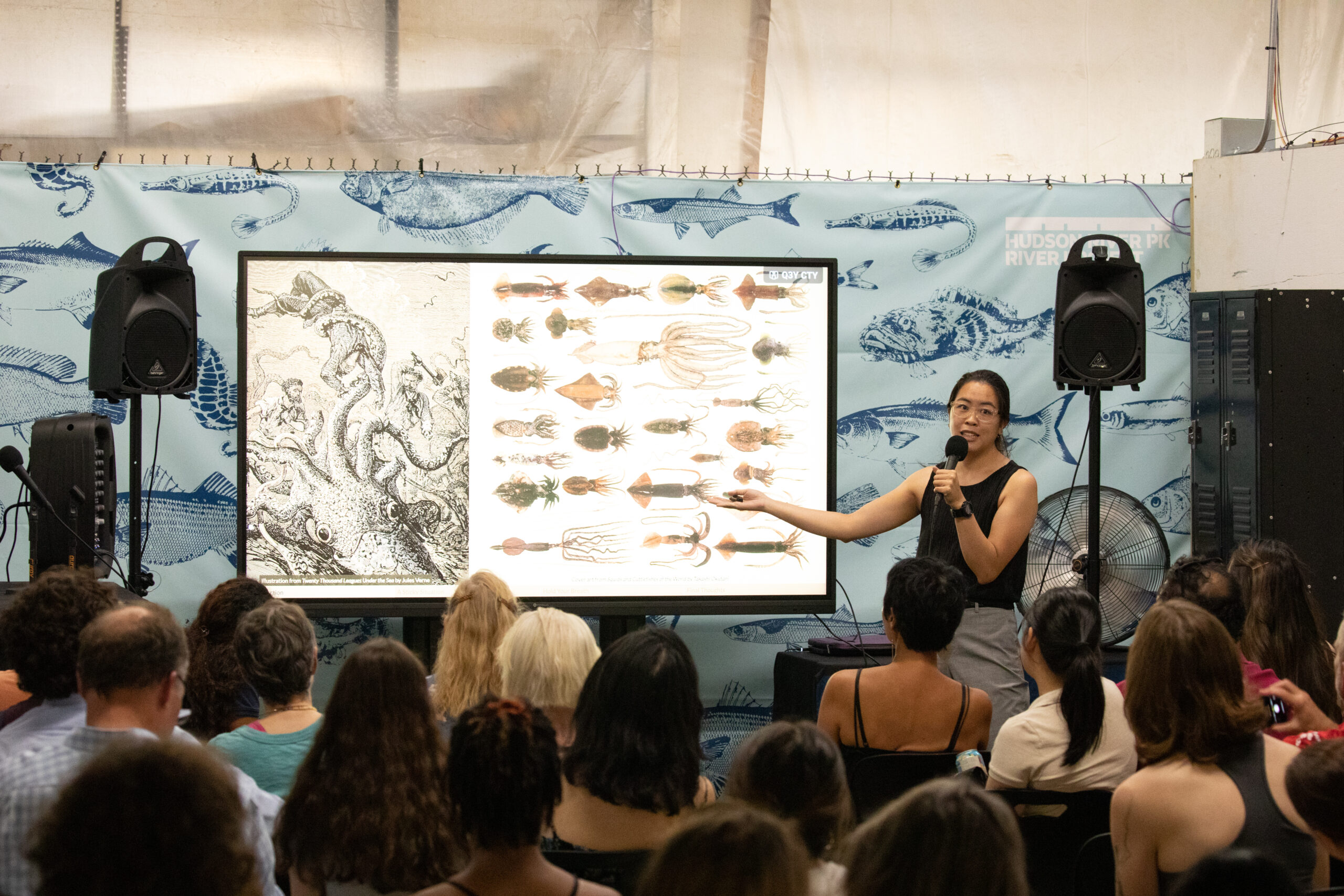 A scientist makes a presentation to a seated audience at the Pier 40 Wetlab during an Ask a Scientist event