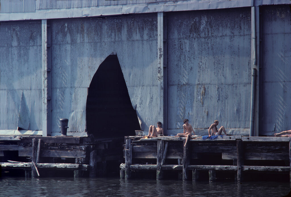 Men sunbathing on a pier. A building behind them has a massive tear in its exterior wall.