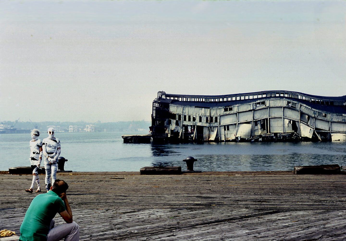 Two people wrapped in bandages like mummies walk on a Hudson River pier. A collapsing pier is visible in the background.