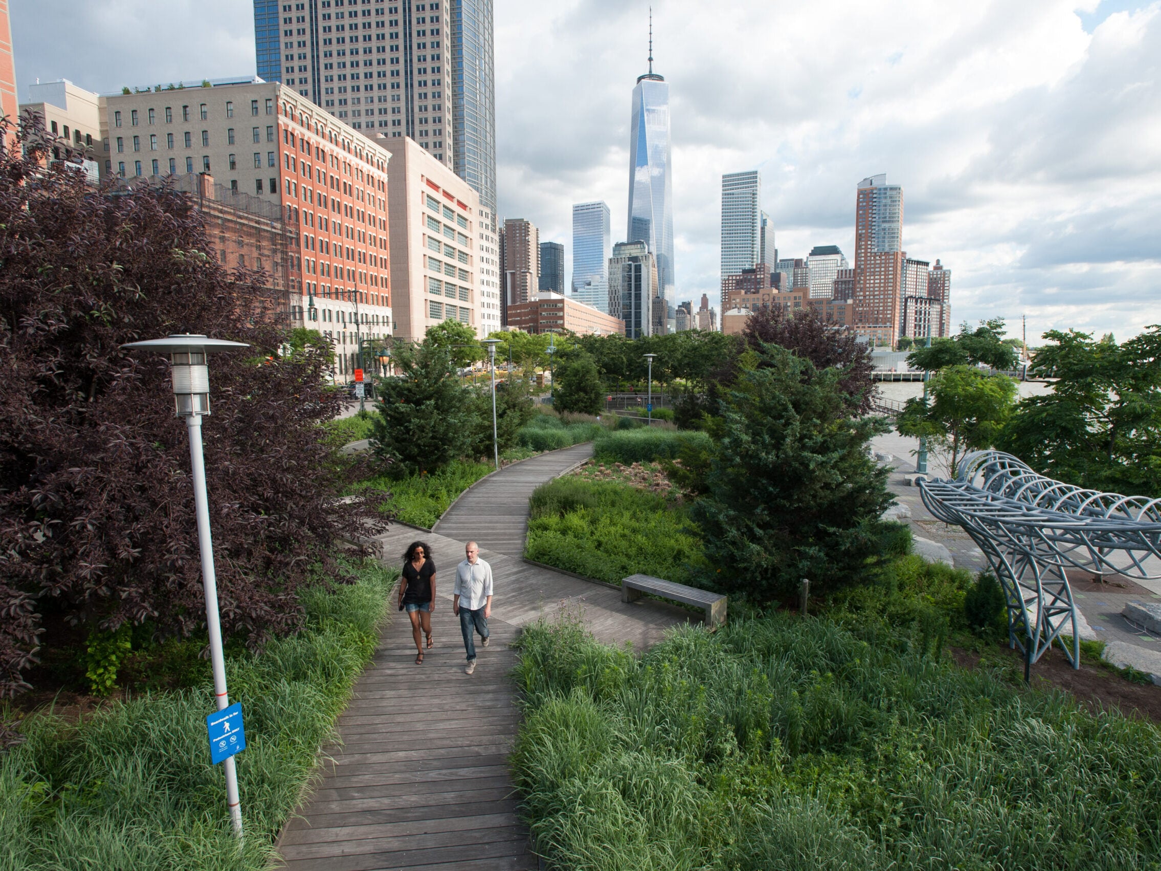 Two people walking on the Tribeca Boardwalk in Hudson River Park. One World Trade Center is visible in the background.