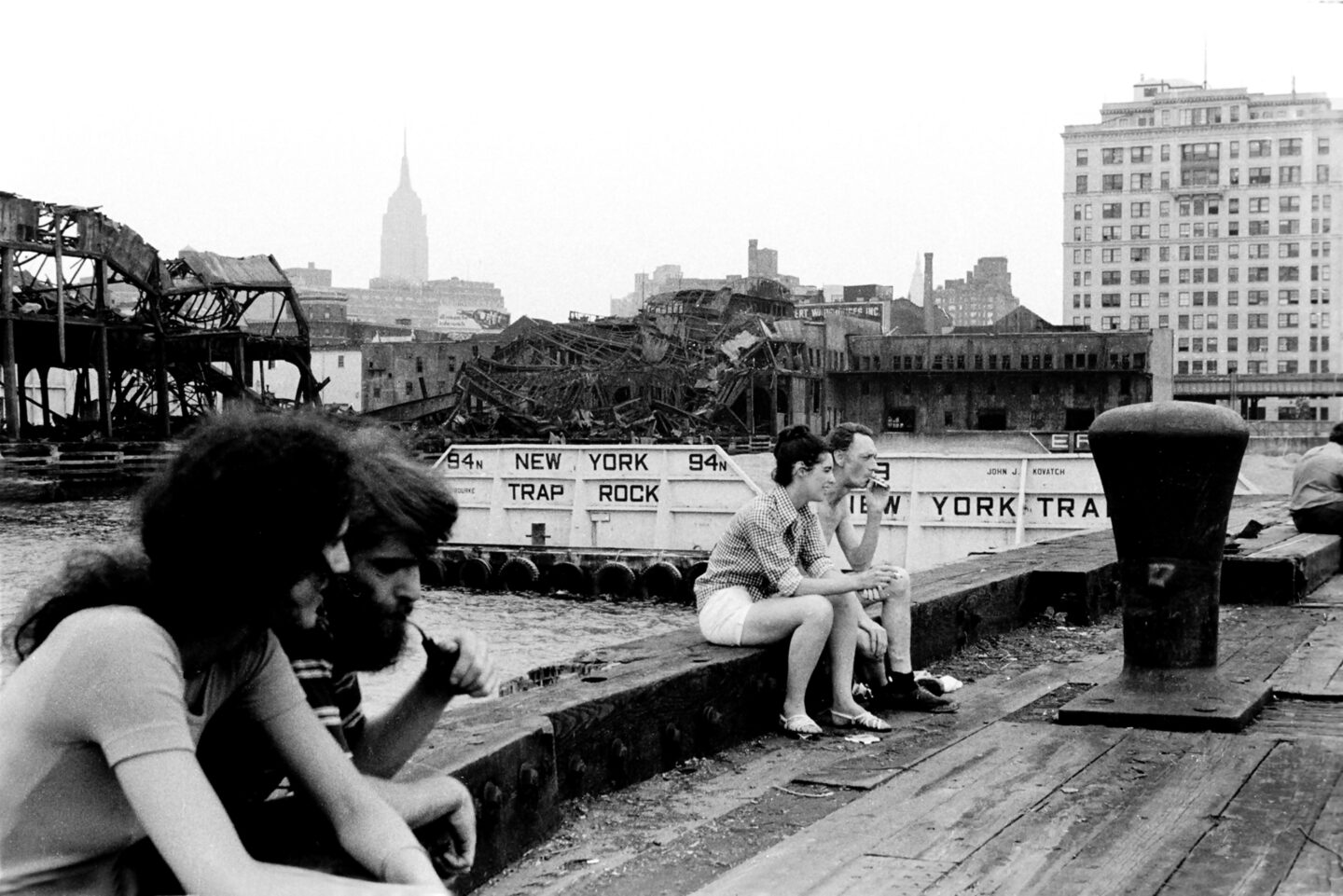 People sitting on the wall of a Hudson River pier. A collapsing pier and the Empire State Building are visible in the background.