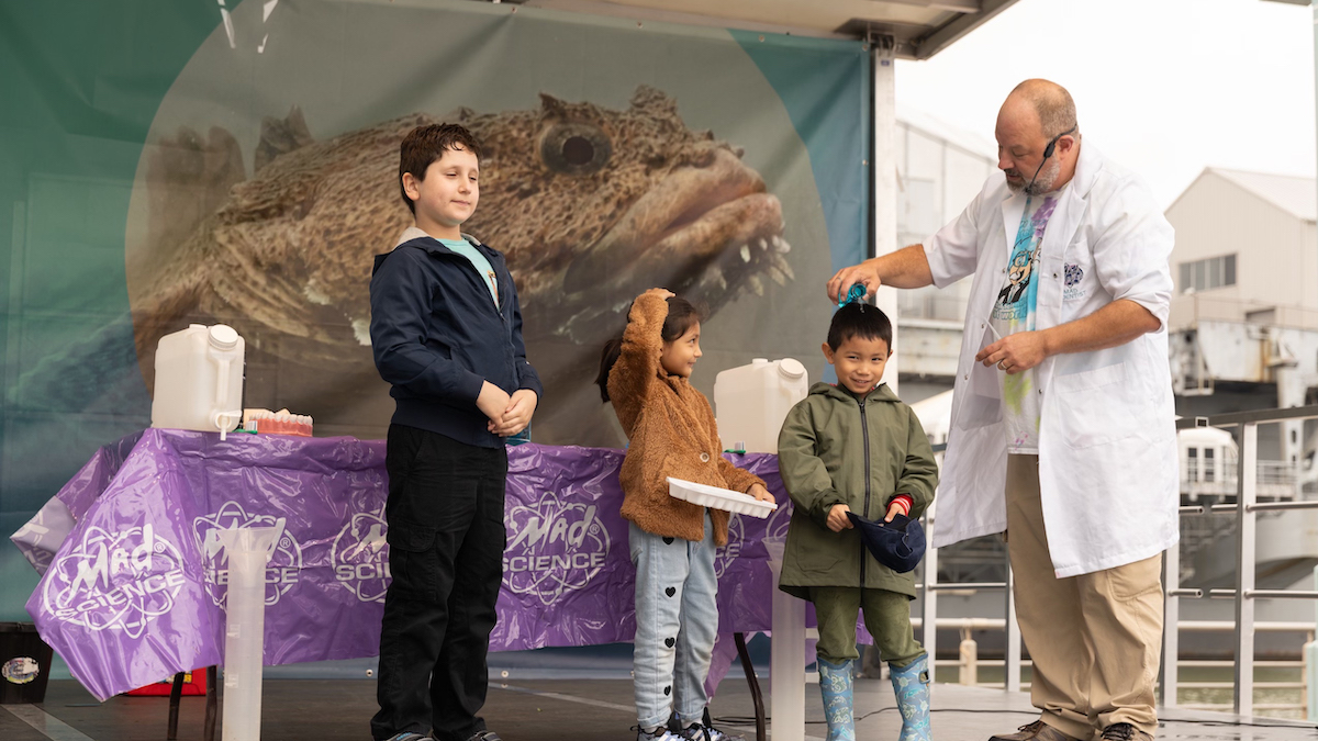 A presenter from Mad Science pours a liquid over a child's head while two children on stage observe