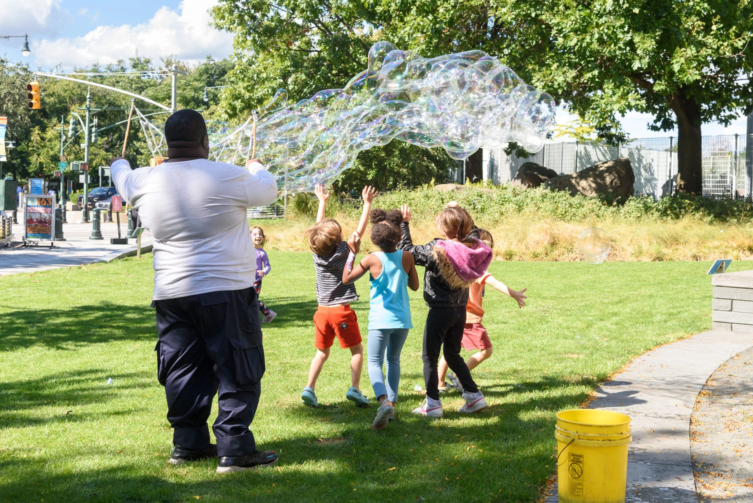 A bubble performer creates a long string of bubbles as children jump and play below them on the Chelsea Waterside lawn