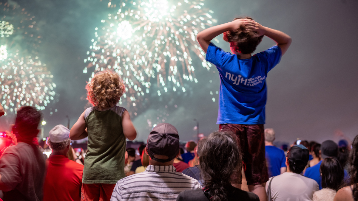 Two children sit atop adults' shoulders in a crowd watching the Macy's 4th of July Fireworks in Hudson River Park