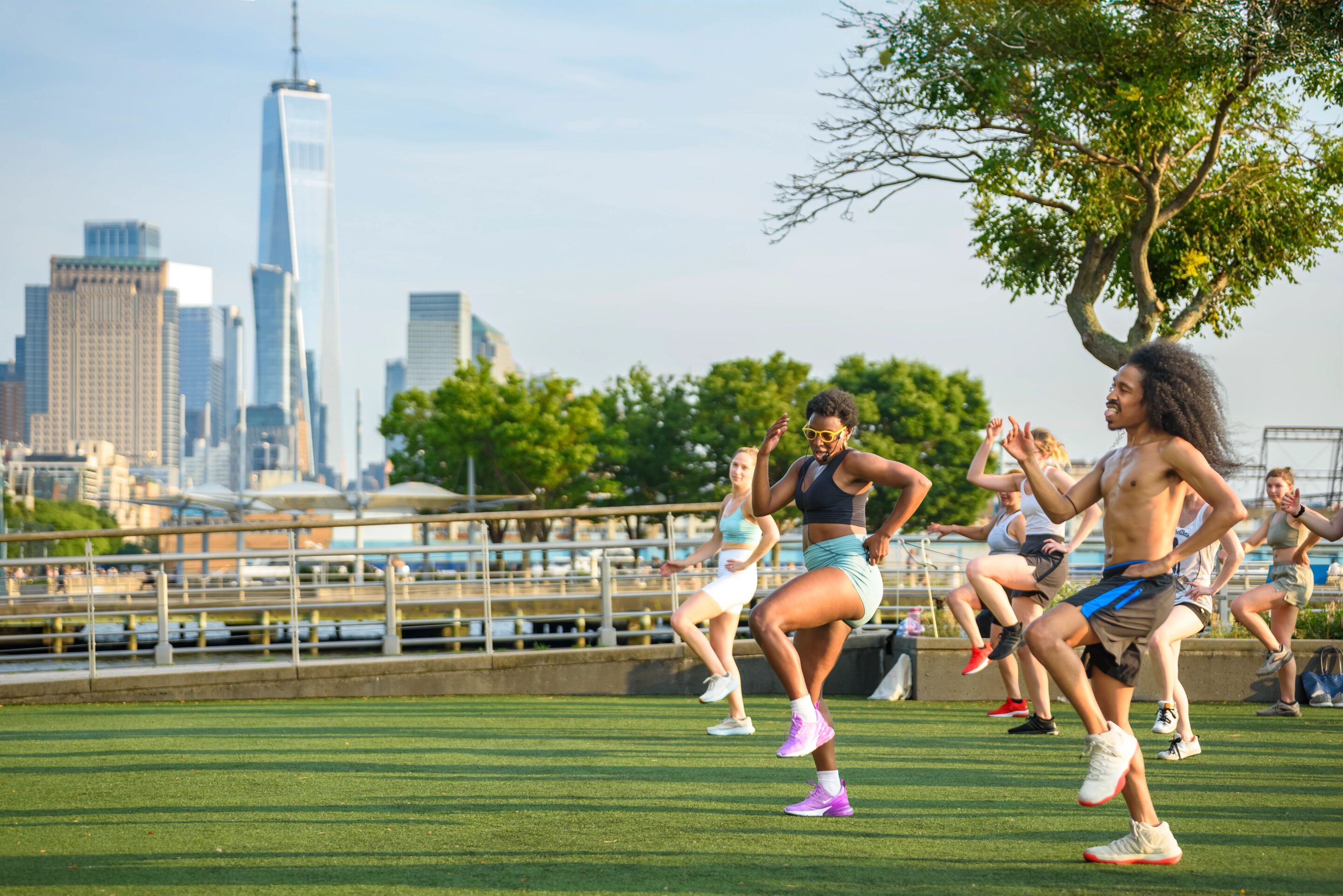 A fitness instructor leads a Healthy on the Hudson class in a "running in place" exercise on the Pier 46 turf field. One World Trade Center is visible in the distance.