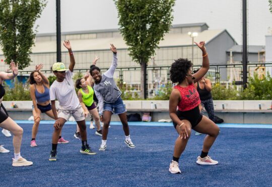 A group fitness instructor leads a class of Healthy on the Hudson participants in a stretch on the Pier 97 Athletic Field