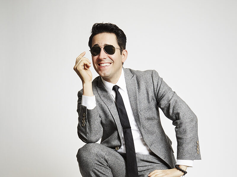 John Lloyd Young sitting on a stool in a gray suit against a white backdrop