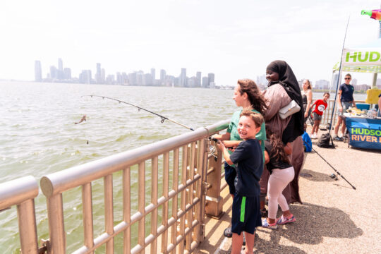 Children fishing from a Hudson River Park pier, with families gathered along the railing.