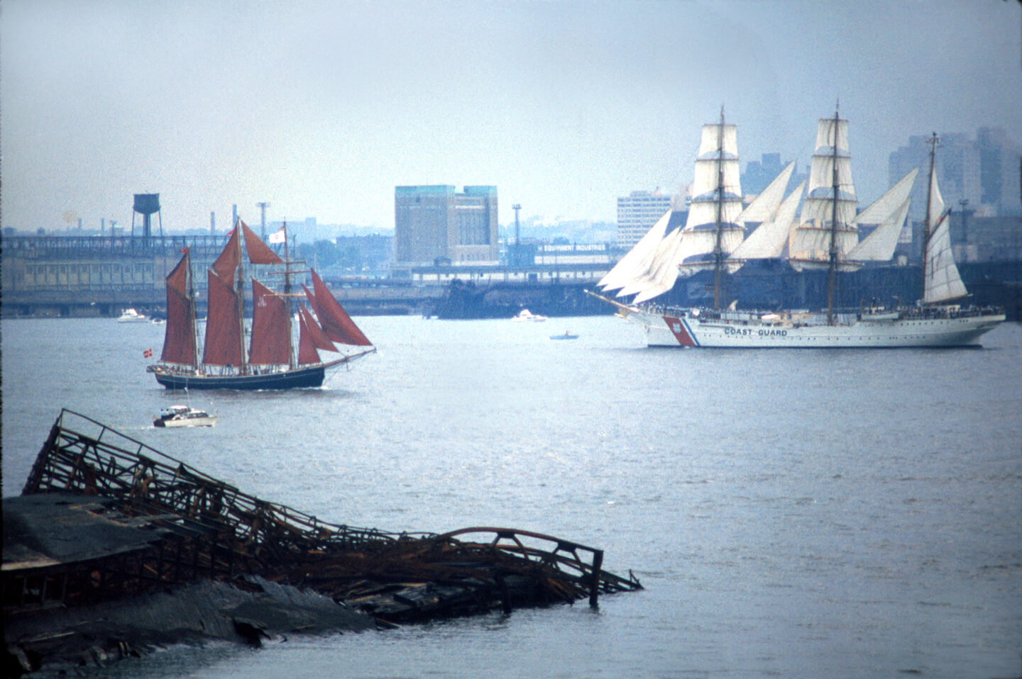 Tall ships sailing on the Hudson River, including a Coast Guard vessel with white sails and a smaller schooner with red sails, with the New York City waterfront in the background.
