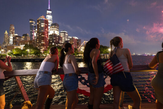 Group of young women gather at Pier 26 at night to watch fireworks over the Hudson River; Lower Manhattan lit up in the background