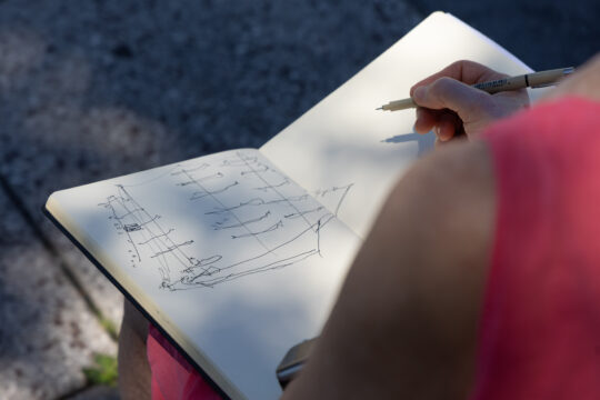 Person in red shirt drawing a ship in notebook