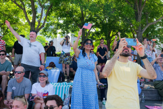 Crowd of people in front of trees; some taking photos, one woman in dress waves American flag