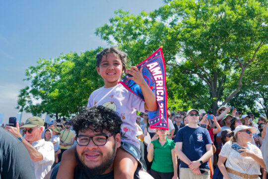 Crowd of people in front of trees, in foreground a young girl sits on a man's shoulders; she is holding an America's 250th flag