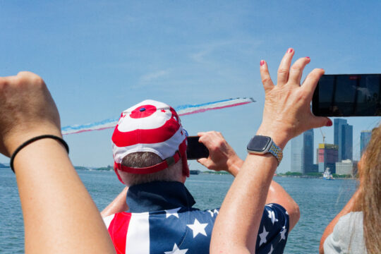 People hold phones up to capture view of planes trailing red white and blue overhead