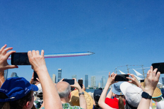 Crowds hold phones up to capture view of planes trailing red white and blue overhead