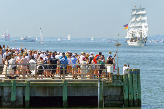 Crowd of people on edge of pier with tall ship in the distance on New York Harbor
