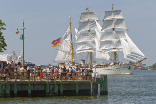 People gather at edge of a pier to watch tall ship sailing Hudson River