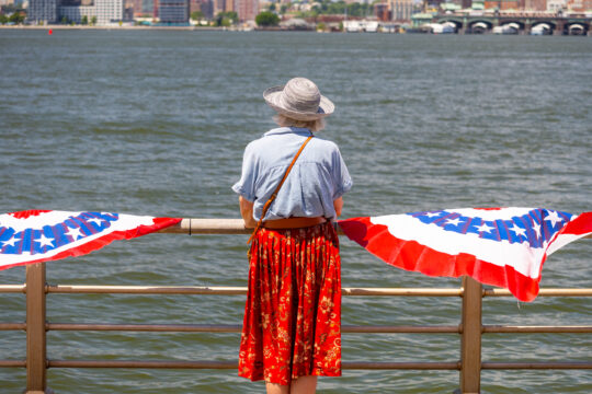 Woman in blue shirt and red skirt stands in front of railings in front of Hudson River; red white and blue hangings on railings