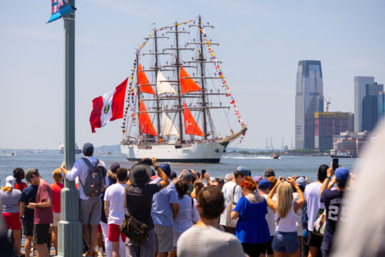 People gather at edge of a pier to watch tall ship with red and white sails sailing Hudson River