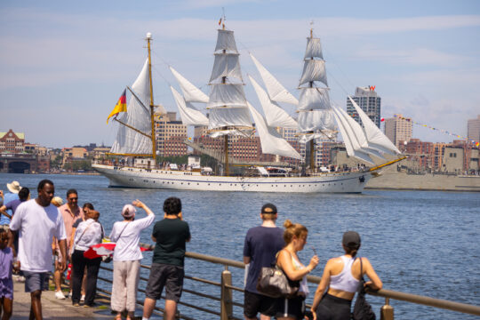 People gather at edge of a pier to watch tall ship sailing Hudson River